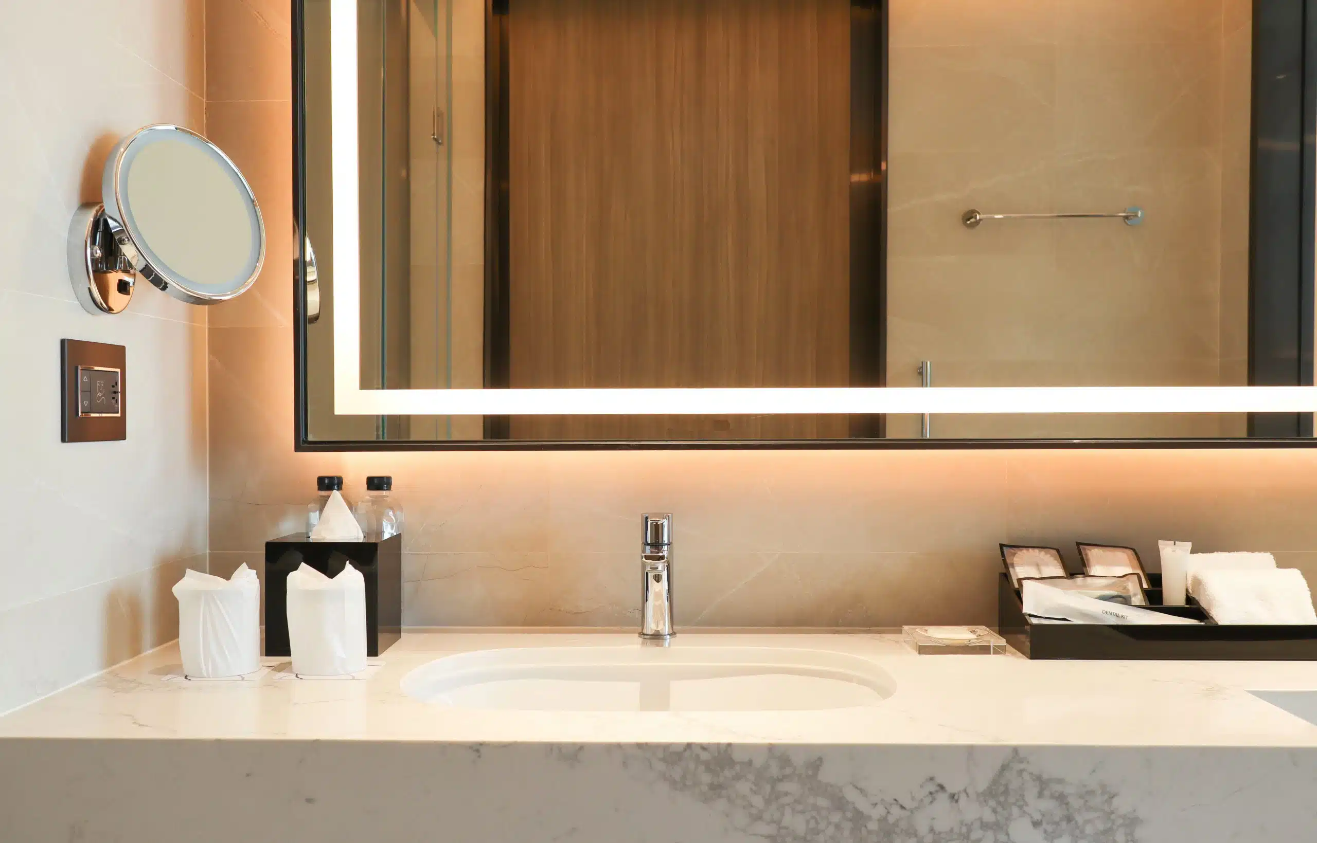 Modern bathroom vanity with marble countertop, integrated oval basin, chrome tap, and a large illuminated LED mirror, complemented by a wall-mounted magnifying mirror and warm ambient lighting