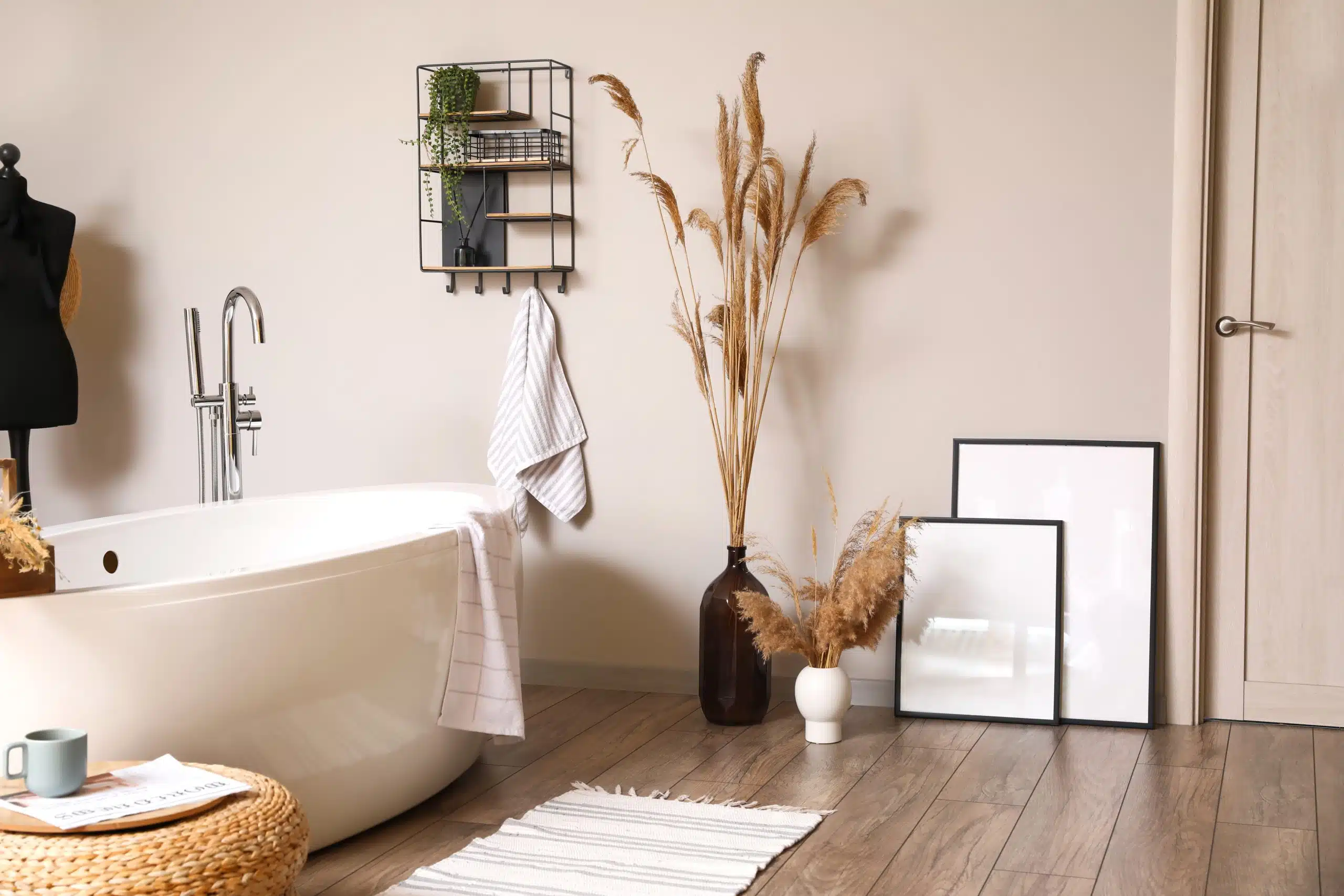 Modern boho-style bathroom featuring a white freestanding bath with chrome floor-mounted tap, wooden flooring, neutral walls, and decorative accents including dried pampas grass in vases, a black wire shelf, framed prints, and soft striped towels